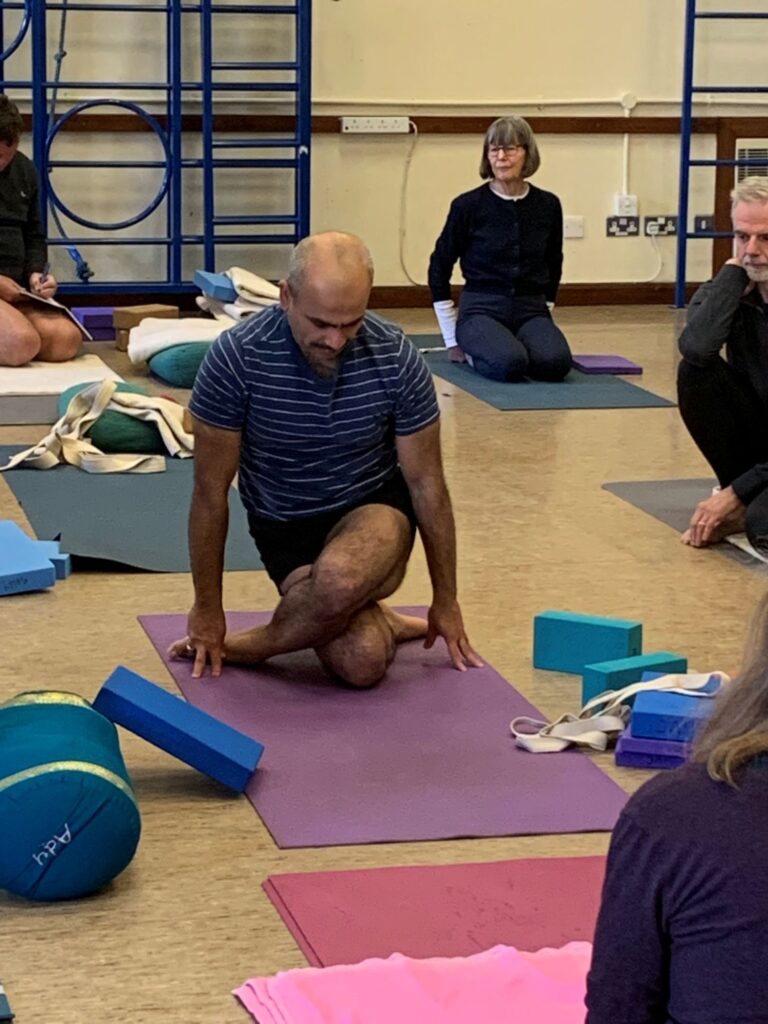 yoga teacher demonstrating pose gomukhasana, watched by people in a gym